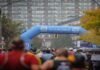 Zone 2 and LT1 Training: Stop Marathon Fade A crowded marathon course looking from behind a sea of runners moving toward a large blue inflatable Abbott archway that reads "life. to the fullest." A race clock stands on the right next to autumn trees, with a large, multi-story apartment building filling the background on an overcast day.