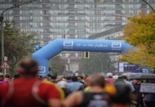 LT1 and Zone 2 Training: Stop Marathon Fade A crowded marathon course looking from behind a sea of runners moving toward a large blue inflatable Abbott archway that reads "life. to the fullest." A race clock stands on the right next to autumn trees, with a large, multi-story apartment building filling the background on an overcast day.