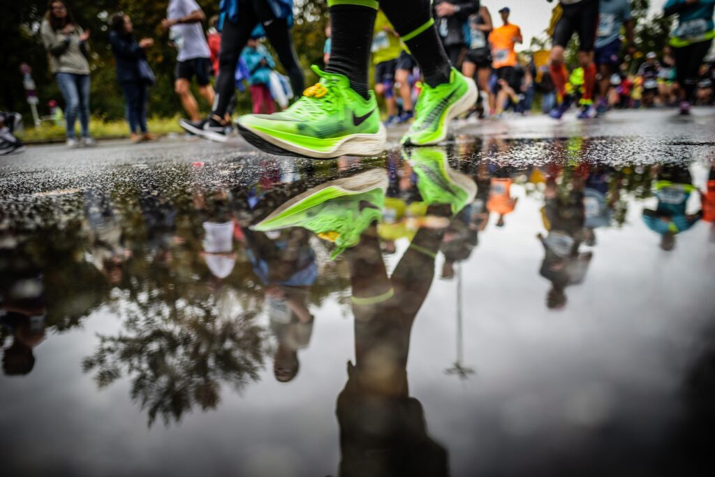 Neon green running shoes mid-stride beside a puddle reflecting runners at a 10K road race